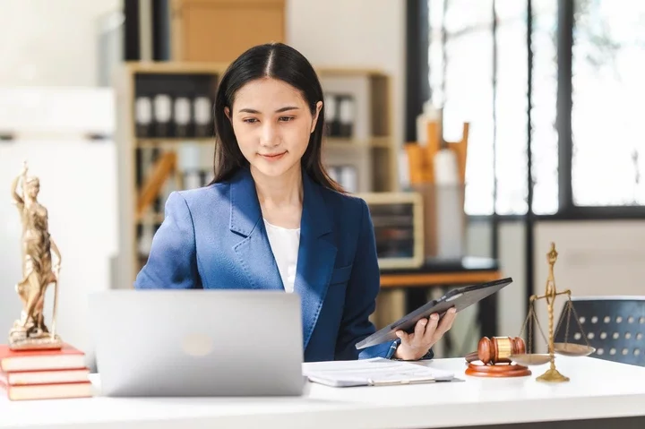 A professional woman in an office holds a tablet, seated at a desk with legal books, a gavel, and scales of justice, indicating a legal profession