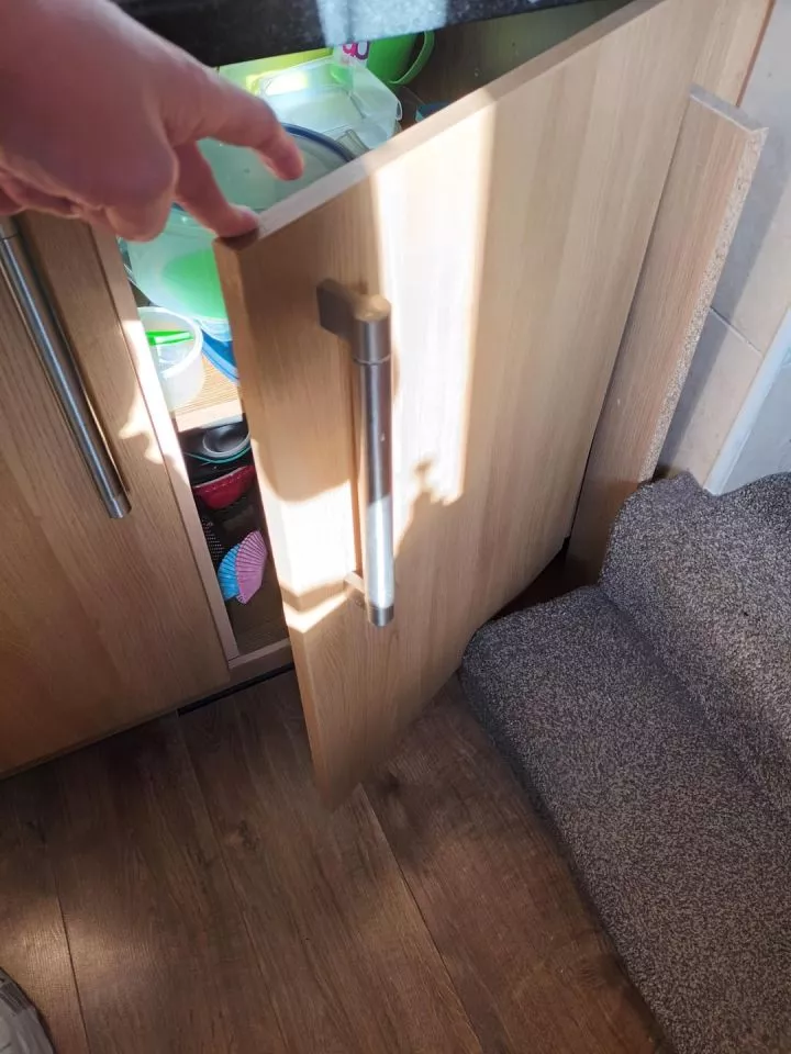 A hand opens a kitchen cabinet door, revealing dishes and utensils inside. The bottom of a staircase blocks door from opening more than 45 degrees
