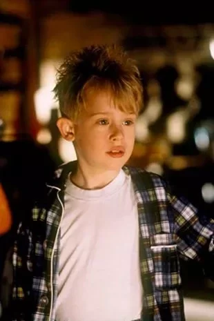 A young Macaulay Culkin as Kevin in an open gingham shirt over a white t-shirt