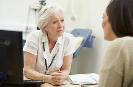 An older woman GP sitting at a desk across from a younger woman