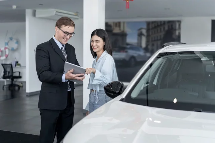 A businessperson in a suit shows a young woman in casual clothes a tablet, standing near a car in a showroom, suggesting a vehicle purchase