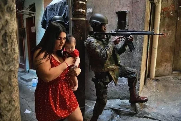 A woman walks with her baby past a PM militarized police soldier in position and aiming his rifle in Rocinha favela in Rio de Janeiro