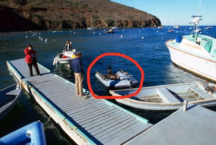 Two people stand on a dock next to small boats in a calm bay with rocky hills in the background