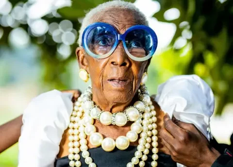 Luxury Media Zambia A  hand can be seen adjusting one of Margret Chola's puffed white sleeves. She is wearing big blue-framed sunglasses and four strings of pearls of various sizes