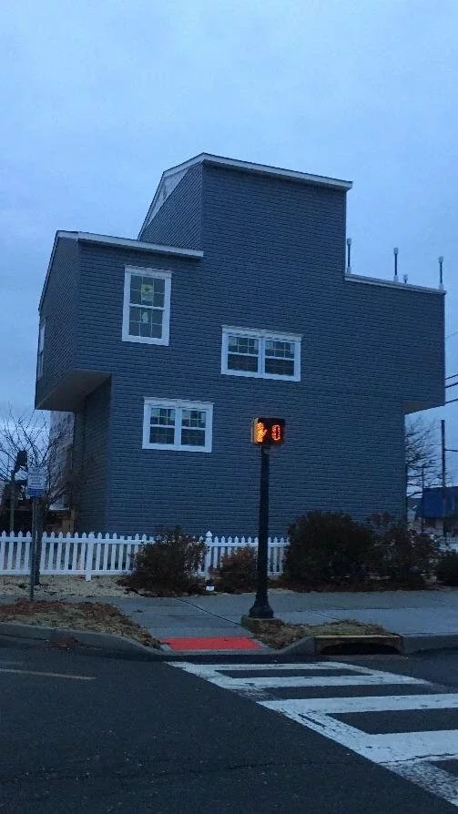 Large, asymmetrical gray house with large windows, next to a white picket fence and crosswalk