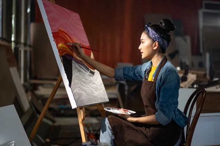 A person in an art studio painting on a canvas; they are wearing a denim shirt and an apron, focusing intently on their work