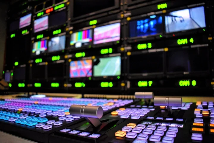 Control room with a digital audio mixer and multiple video screens displaying a variety of channel feeds, suggesting media production work