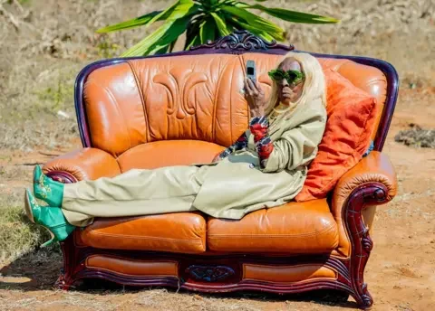 Luxury Media Zambia Margret Chola lying on a orange leather sofa in a garden looking at her phone. She has on a wig of blond hair and is wearing a pale green leather loose-fitting trouser suit with green high-heeled boots and green-rimmed sunglasses 