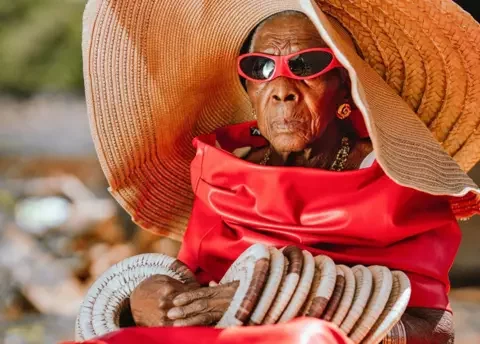 Luxury Media Zambia A seated Margret Chola looks serious as she wears a giant straw hat with bangles that look like straw mat up both her arms. She is wearing red sunglasses and what looks like a red leather dress