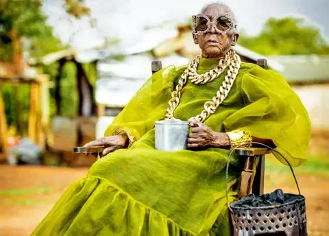 Luxury Media Zambia A seated Margret Chola wearing a chunky gold chain over a green taffeta like dress with puffy sleeves and a big frill for the hem holds a tin mug in her hand. Hanging off the arm of her chair is a 'mbaula