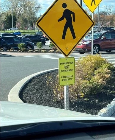 Pedestrian sign at a road. Small sign below reads: &quot;Intersection NOT roundabout. Proceed as 4-way stop&quot;