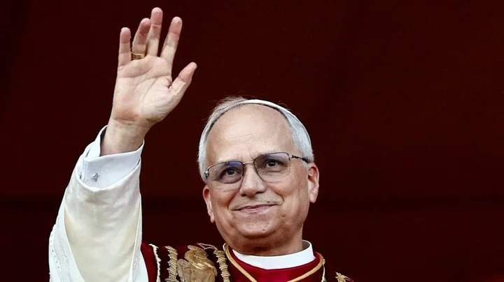 Pope Leo, wearing glasses and religious dress, waving against a red background