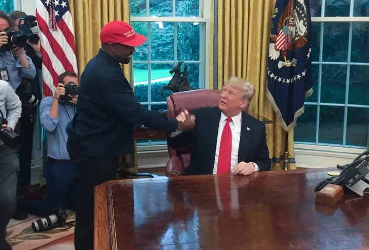 Kanye West wears a red hat and shakes hands with Donald Trump, how sits at a desk.