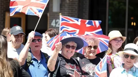 AFP/Getty Images Well wishers wave flags before the arrival of Britain's King Charles III and Queen Camilla at a Lansdowne Park community event in Ottawa, Canada, May 26, 2025. 