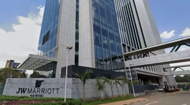 A modern hotel with a grey exterior with lots of floor to ceiling windows down the side of the building. A sign outside says JW Marriott Nairobi. It has some greenery in front of it. 