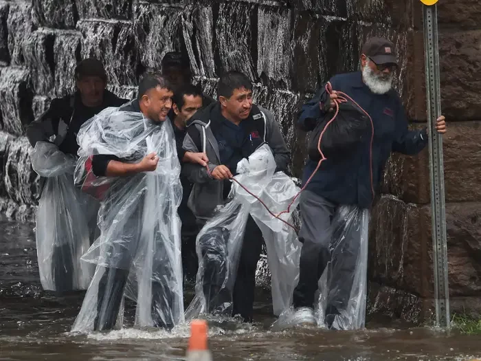 A group of people in clear plastic wrap knee deep in rain water emerge from a tunnel