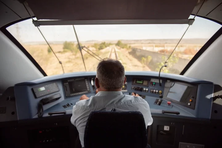 Train engineer operates controls in locomotive cabin, focusing on tracks ahead, illustrating work in the rail transport industry