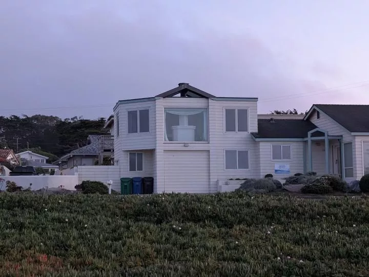 Two-story house in a coastal neighborhood with a large bay window, revealing a fireplace centered inside