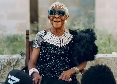 Kooma Jnr A seated Margret Chola laughs at the camera. She is wearing an Indian-style  head band, big bejewelled green-framed sunglasses, a diamond-like waterfall necklace and a black feather boa over a top covered in sequins