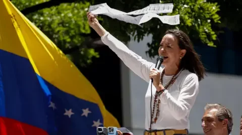 Reuters Venezuelan opposition leader Maria Corina Machado waves a long string of paper in front of a flag as she speaks during a rally.