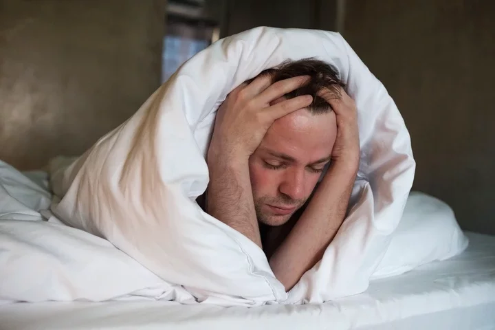 Depressed man lying in his bed wrapped in a duvet with his hands on his head