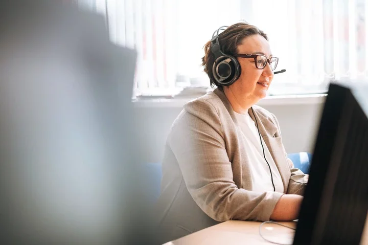 Person in an office wearing headphones with microphone, working at a computer, dressed in a business-casual blazer