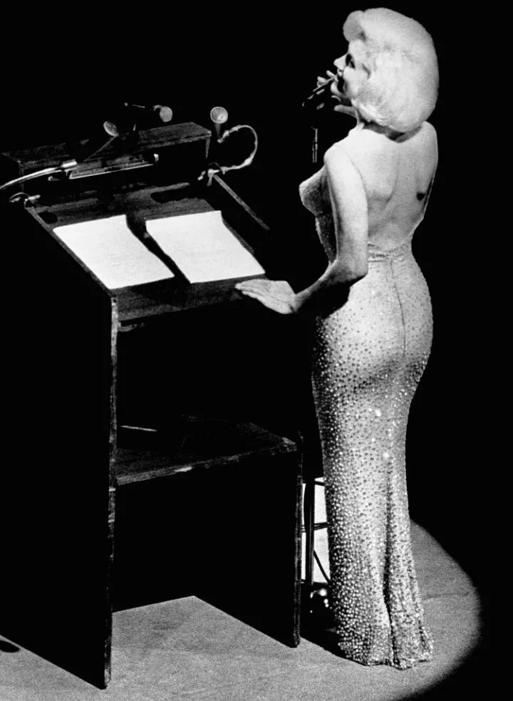 A person in an elegant, sparkling gown stands at a podium, addressing an audience