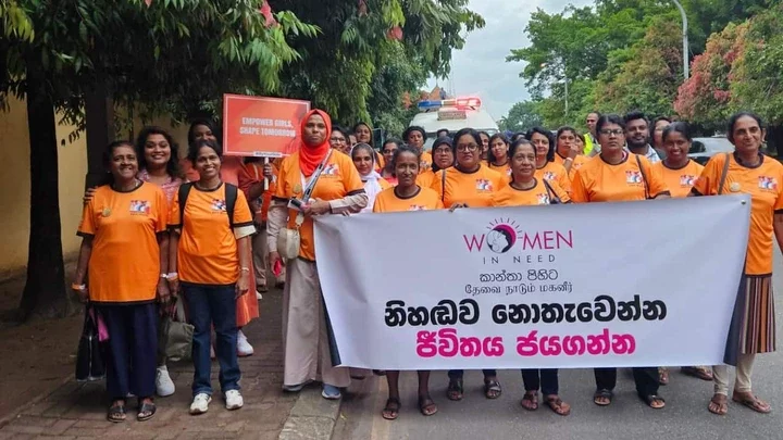 A large group of women wearing orange t-shirts poses with a banner with text in Sinhala language and the words Women In Need' in English at the top.