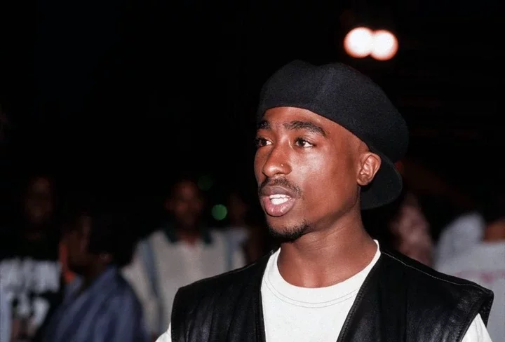 Tupac Shakur wearing a black durag and leather vest speaks with mouth open