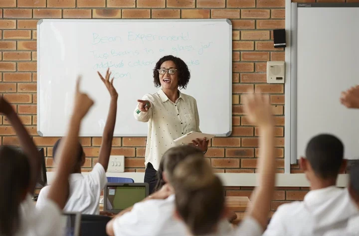 A teacher in a classroom engages with students raising their hands, standing in front of a whiteboard with notes on bean experiments