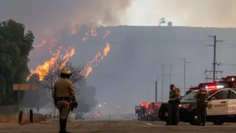 Getty Images Emergency crews patrol along a street as flames burn and smoke billows into the air in the distance