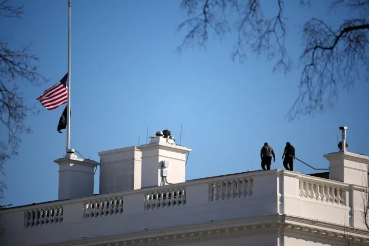 The White House with the American flag at half-staff. Two people are on the roof.